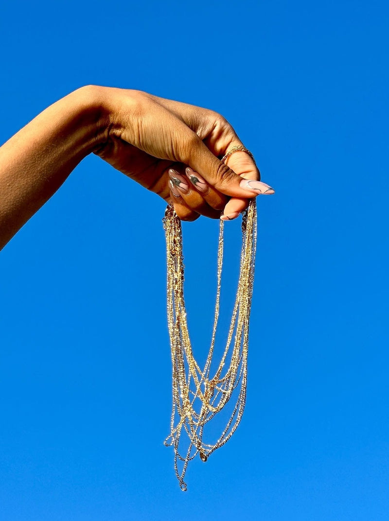 Woman's hand holding a stack of 14K yellow gold Figaro chains with a blue sky background
